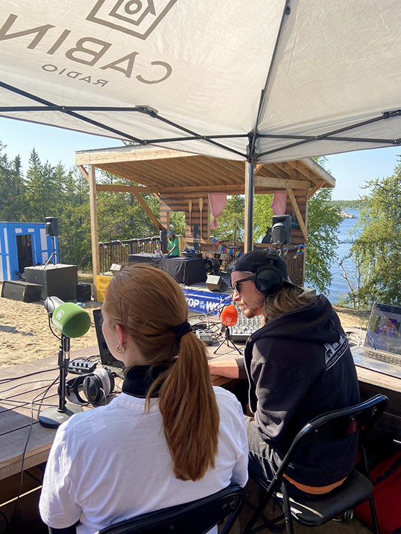 Catriona Koenig (left) sits next to Scott Letkeman (right) broadcasting live from the Cabin Radio lakefront stage. July 2021.